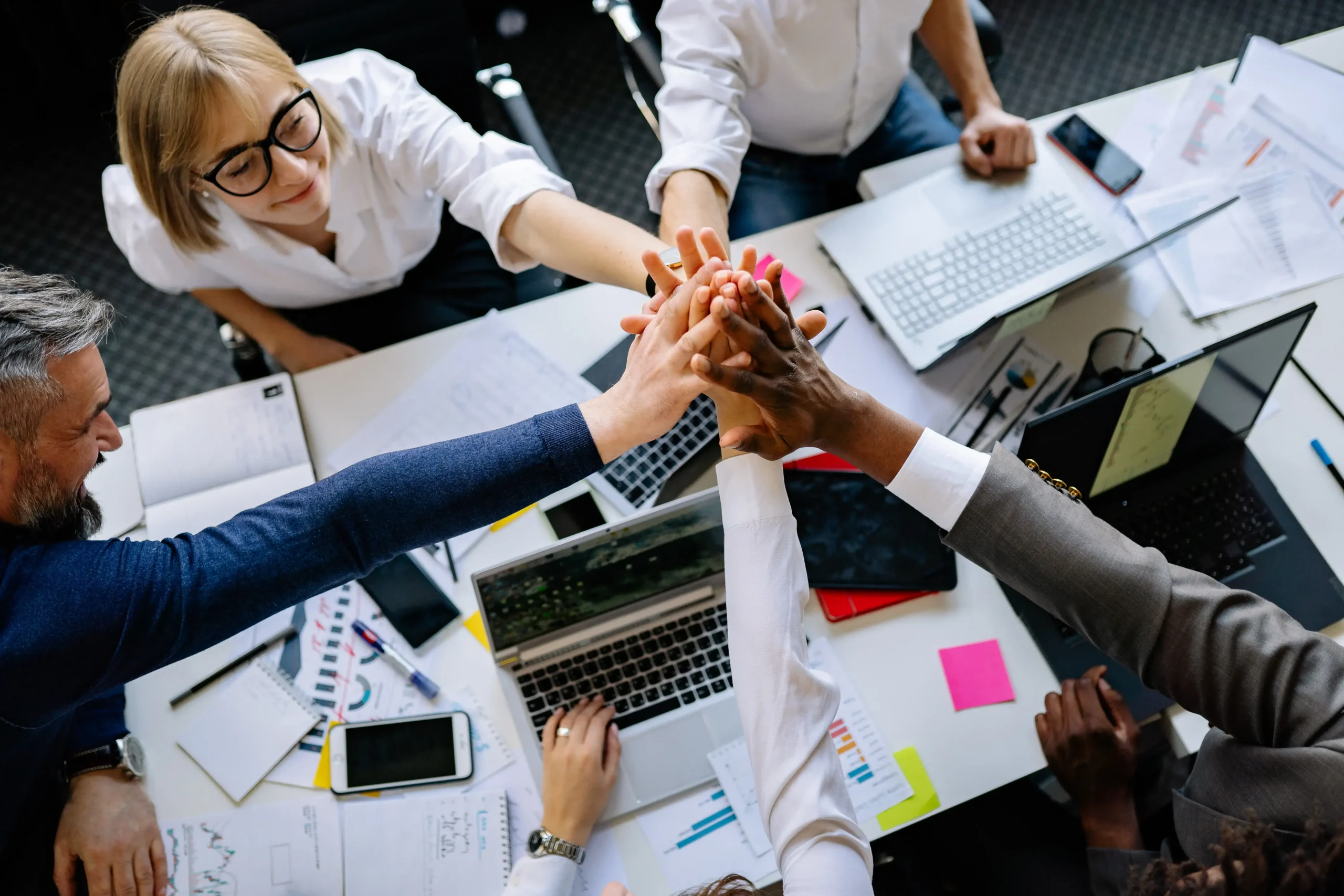Equipo de trabajo celebrando con un gesto de manos unidas sobre una mesa con laptops, gráficos y documentos